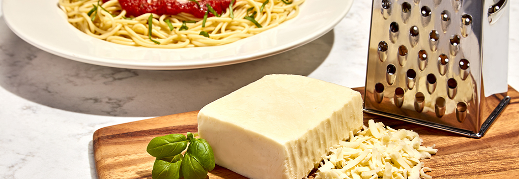 block of white cheese with grater and shreds sitting on a wooden cutting board and a plate of spaghetti and red sauce in the background