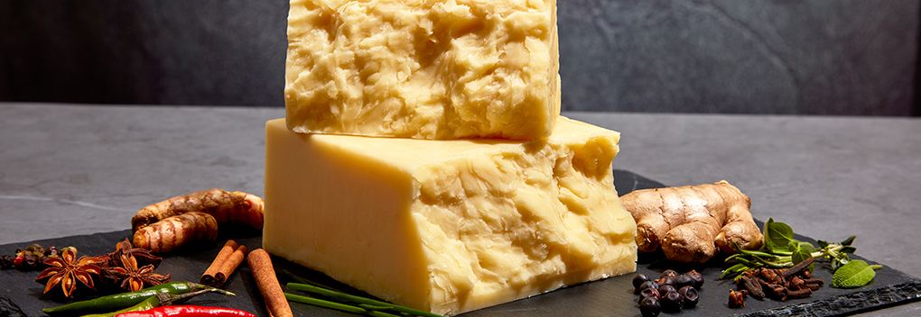 large block of aged, lightly colored cheese split into two halves sitting on a slate board, accented with hot peppers and a variety of spices