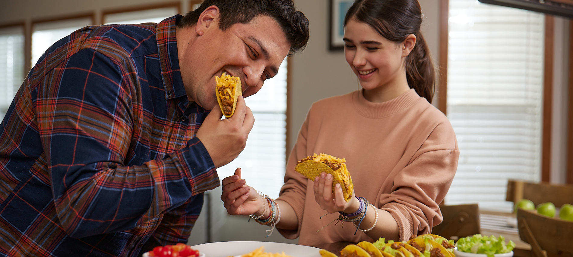 father and daughter eating tacos together in their kitchen, with taco ingredients and cheese prepared on the counter