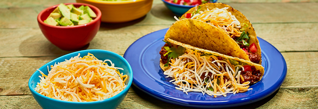 hard shell tacos and ingredients including avocado and shredded cheese displayed in colorful dishes on a table top
