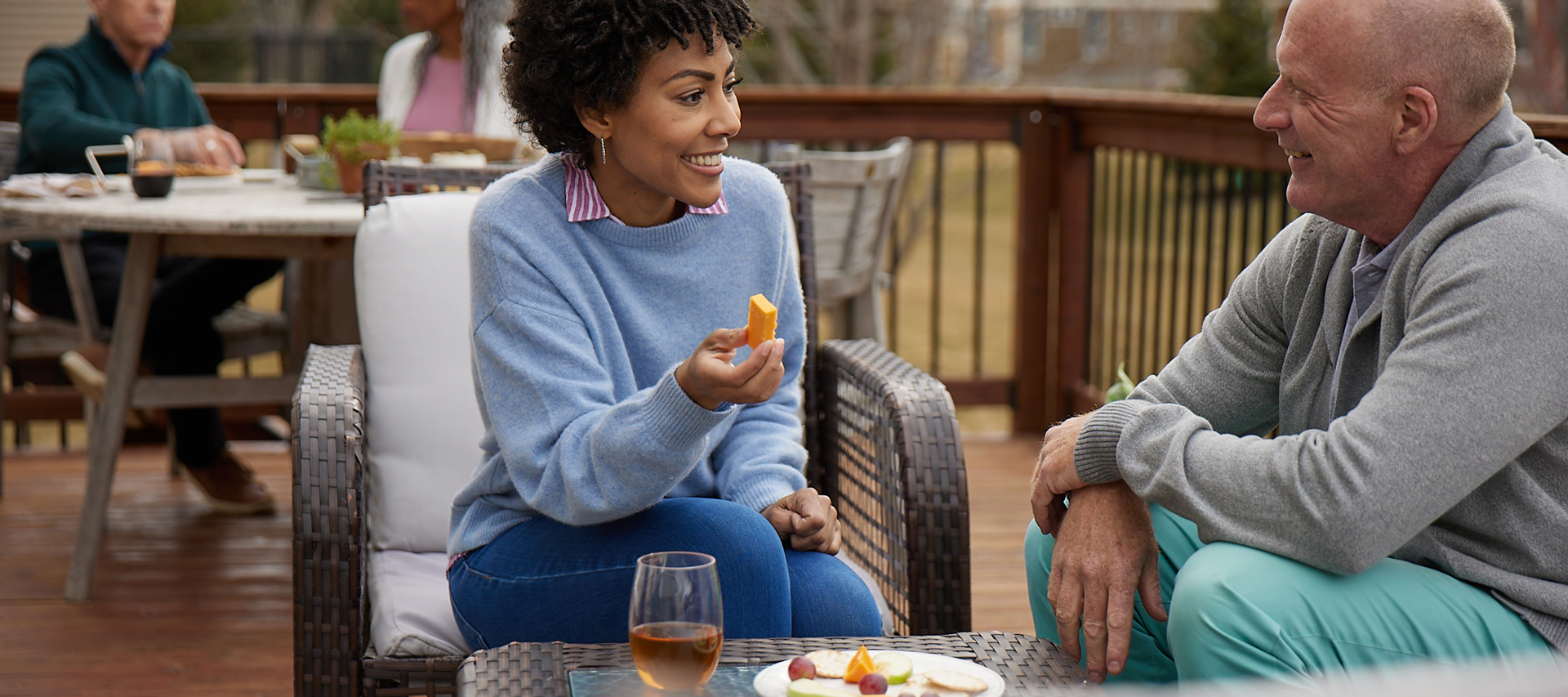 woman eating a piece of yellow cheese while conversing with a man at an outdoor gathering on a patio, you can see a drink and cheese plate sitting on the table in front of them with more people socializing behind them