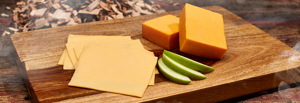 slices of cheese and two cheese blocks sitting on a wooden cutting board next to apple slices with wood chip smoldering in the background