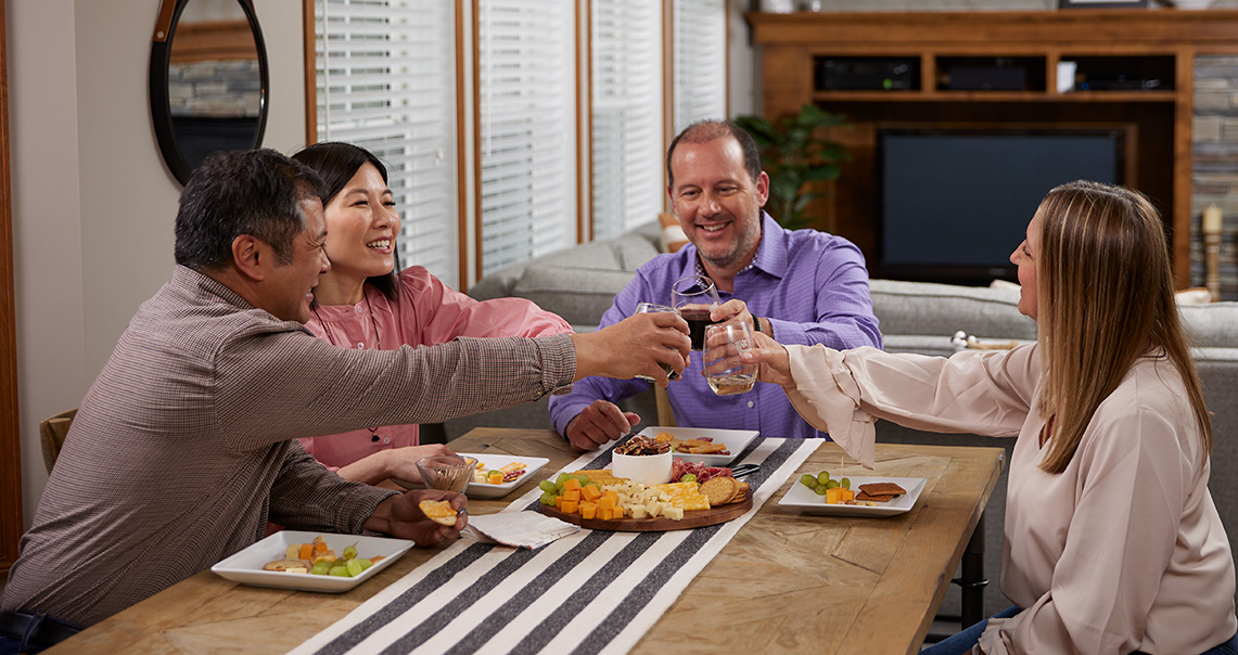 four middle aged adults toast around a table over a charcuterie board of all natural cheeses, grapes and cracker