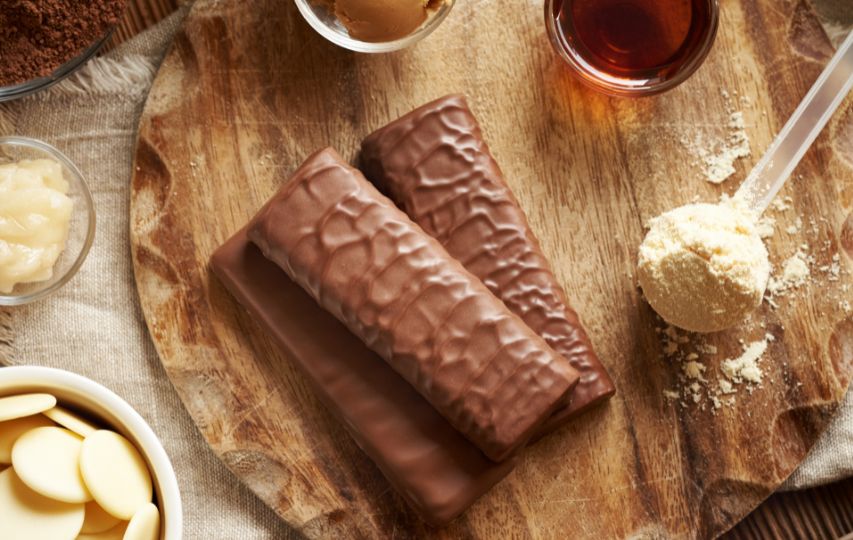 three chocolate covered protein bars stacked on a wooden cutting board next to a scoop of protein powder and other bar ingredients